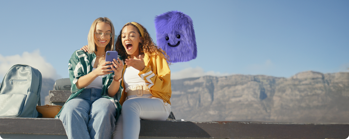 Zwei junge Frauen sitzen im Freien, überrascht beim Handytarif-Vergleich auf ihrem Handy.